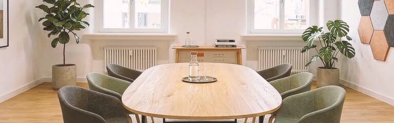 Moderner Besprechungsraum mit großem Holztisch, acht gepolsterten Stühlen, Pflanzen, zwei Fenstern und hellem Tageslicht.