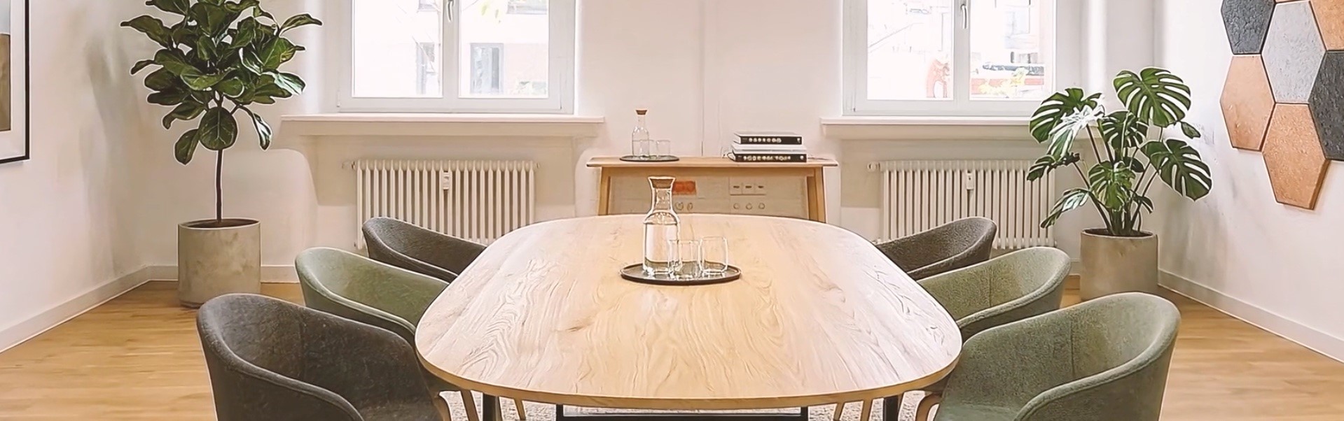 Moderner Besprechungsraum mit großem Holztisch, acht gepolsterten Stühlen, Pflanzen, zwei Fenstern und hellem Tageslicht.