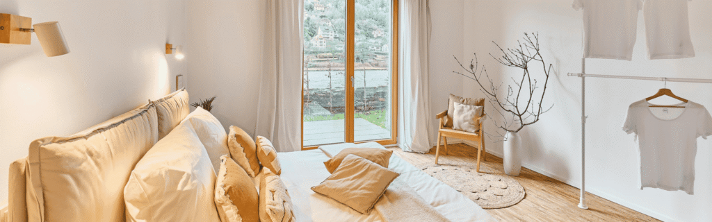 Hell eingerichtetes, minimalistisches Schlafzimmer im Japandi-Stil mit einem großen Bett in Naturtönen, einer großen Fensterfront mit Blick ins Grüne und einem minimalistischen Kleiderständer.
