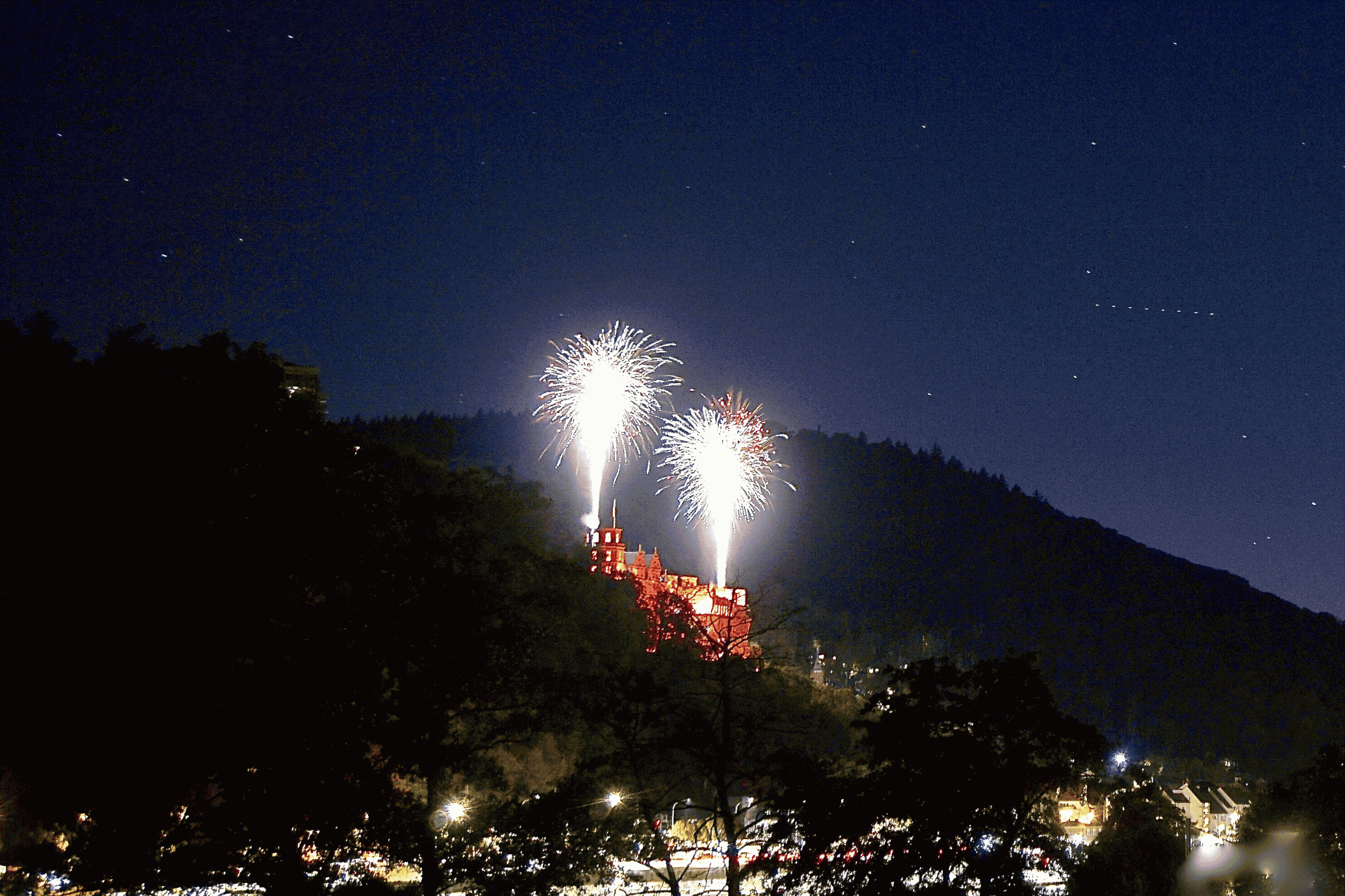 Nächtliche Feuerwerkskörper erleuchten den Himmel über einem Schloss auf einem bewaldeten Hügel. Im Vordergrund sind dunkle Bäume und vereinzelte Lichter zu sehen.